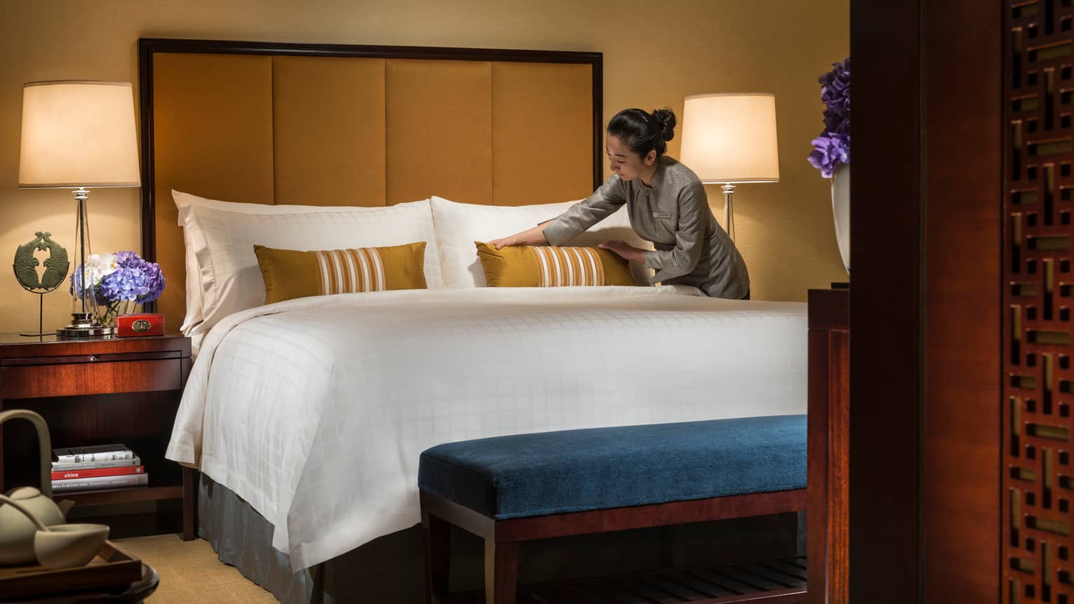 Hotel staff makes bed, adjusts gold accent pillow on white pillows, linens