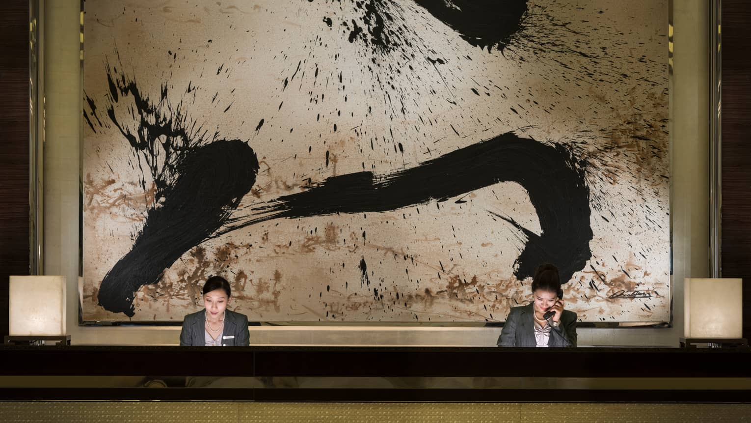 Two hotel reception staff at long desk under large black-and-white abstract painting
