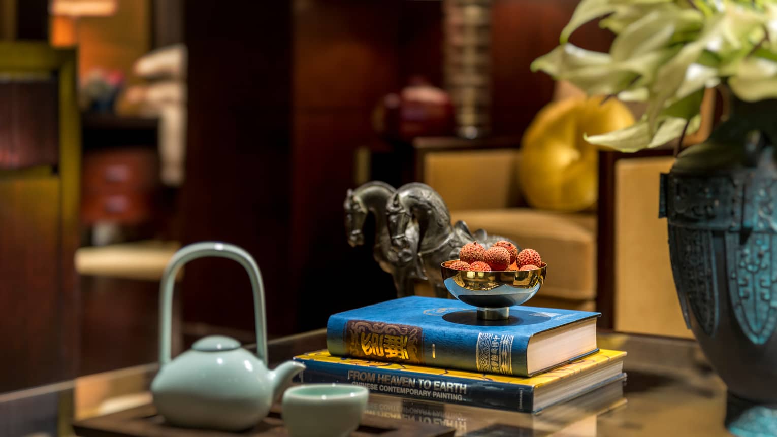 Close-up of white teapot, cup, stack of books, bowl of fruit, horse statues