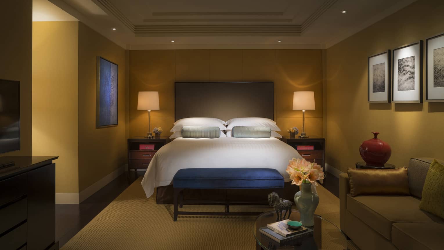 Dimly-lit hotel room bed with tall padded headboard, small blue bench at foot, small table with vase, horse statue