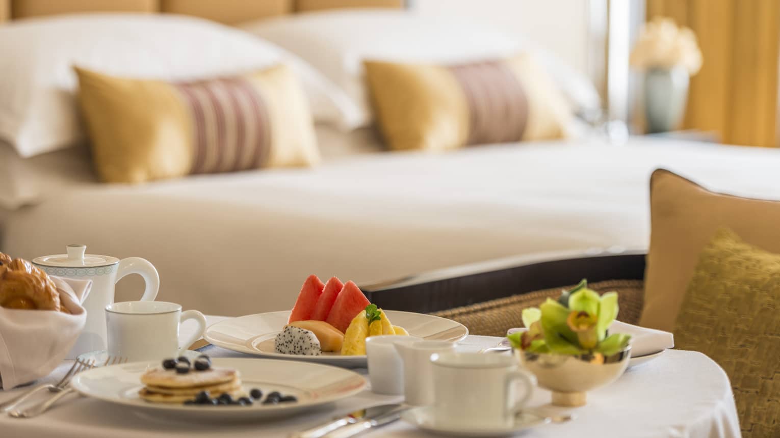 Pancakes, coffee, fresh fruit breakfast items on in-room dining table in front of bed