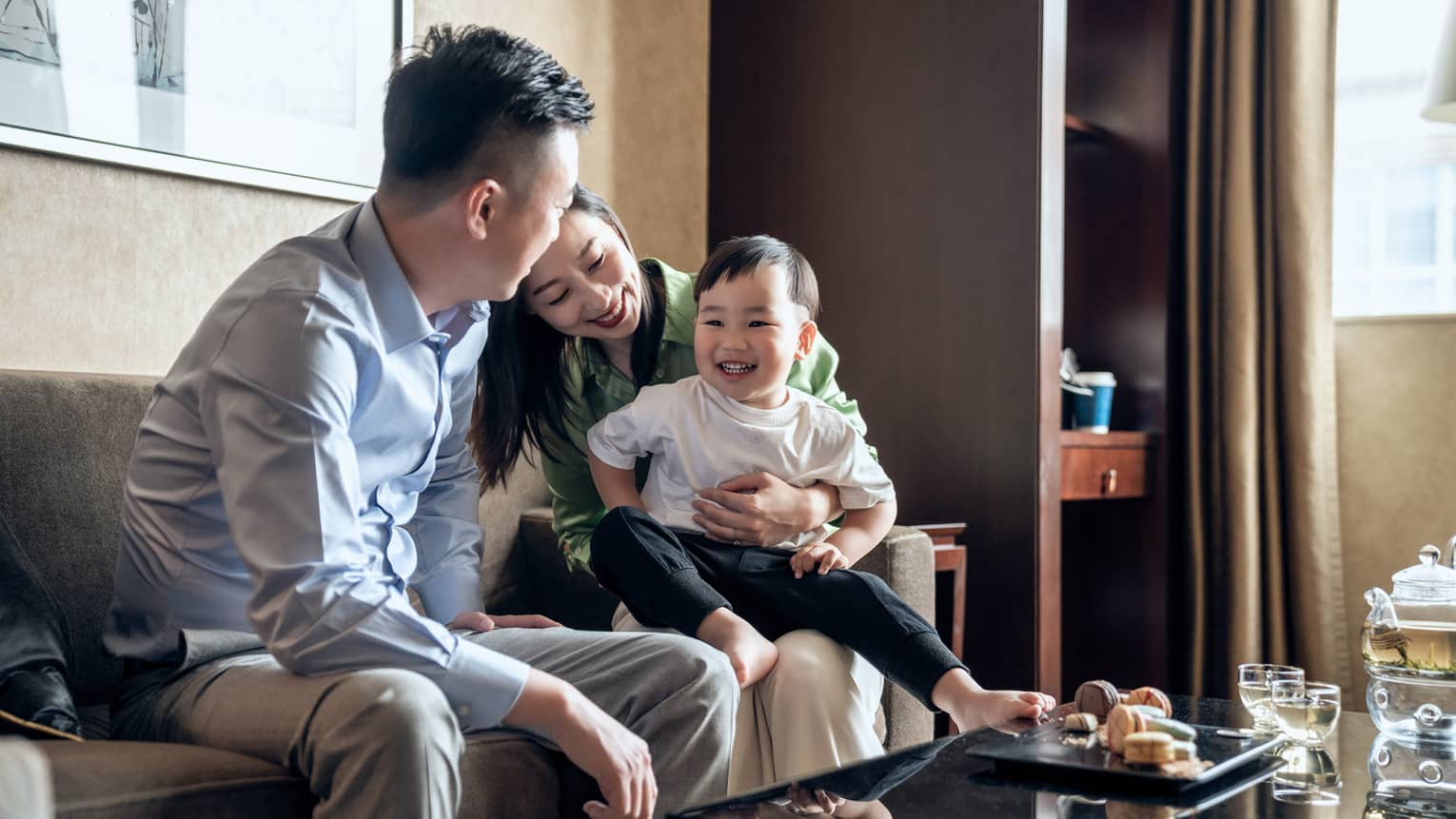 Laughing couple sits with their toddler son, tea and sweets on the coffee table in front of them