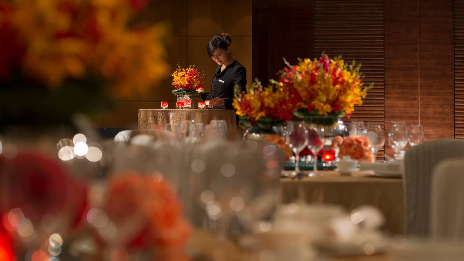 Hotel server in uniform places candles on table with orange flowers, linens, behind dining tables