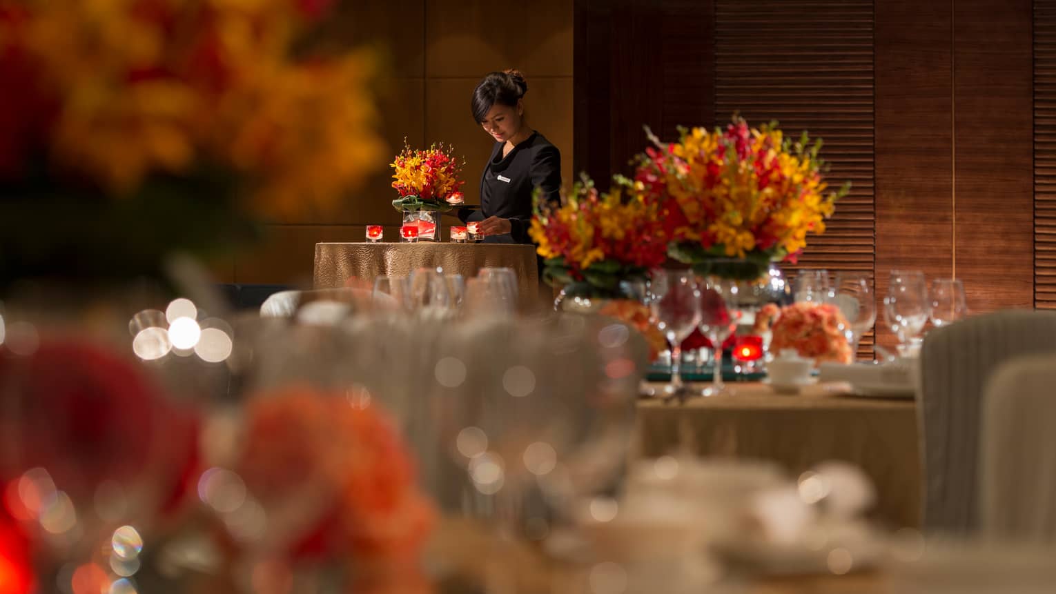 Hotel server in uniform places candles on table with orange flowers, linens, behind dining tables