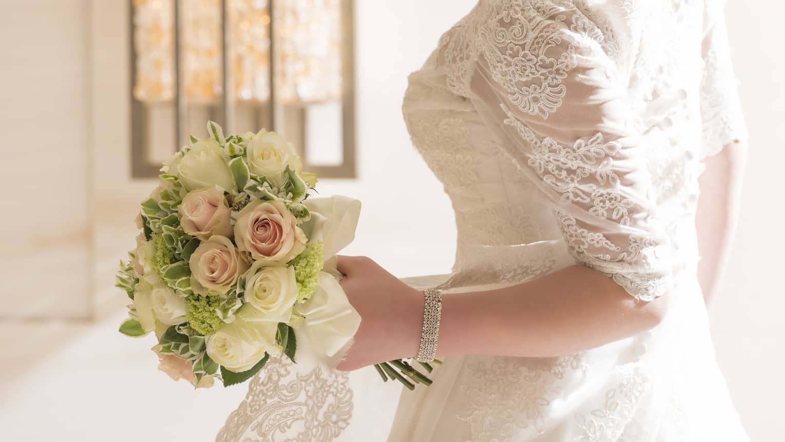 Bride wearing lace wedding gown stands by sunny window, looks down at rose bouquet