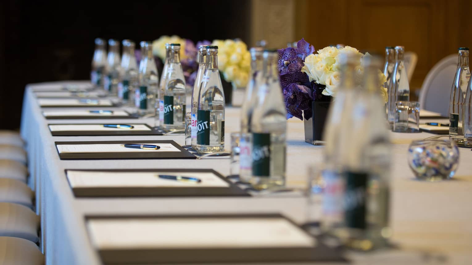 Row of meeting agendas, glass bottles, small vases with flowers on boardroom table